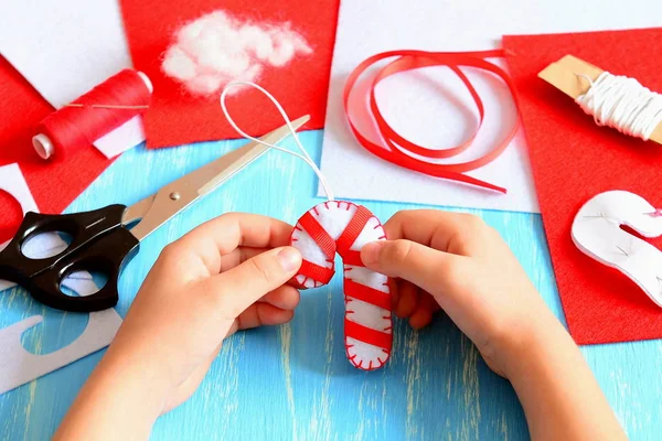 Ein kleines Kind hält den weihnachtlichen Zuckerrohrschmuck in den Händen. Nähmaterial für die Herstellung von Christbaumschmuck. Leichtes und lustiges Papierbasteln für Kinder im Winter. Kinder basteln Zuckerstangen zu Weihnachten. Weihnachtsidee für Kinder — Stockfoto