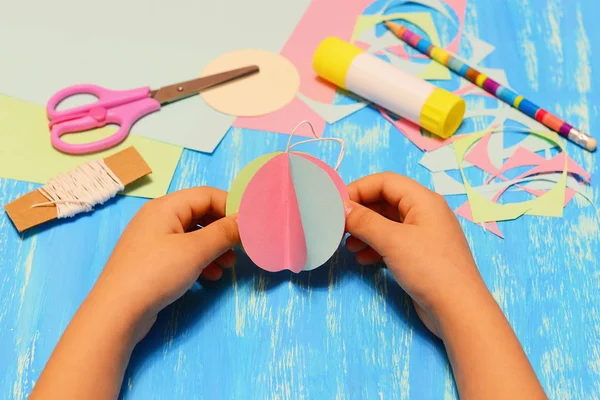 El chico tiene un adorno de bola de árbol de Navidad de papel en sus manos. El chico muestra manualidades de papel de Navidad. Tijeras, lápiz, pegamento, trozos de papel de colores y sobras sobre fondo de madera azul. Artesanía de vacaciones para niños. Fondo de artesanías navideñas —  Fotos de Stock