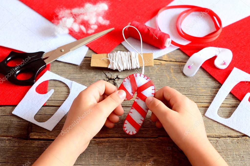 Child holds Christmas felt candy cane ornament in his hands. Materials and tools to create Christmas tree decorations. Simple and beautiful handicrafts for kids to make concept. Kids Christmas symbol DIY idea. Vintage style