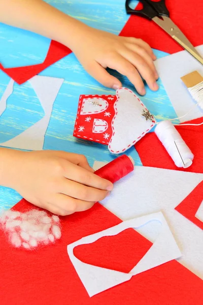 A gyerek karácsonyi kézművességet készít. A gyerek rátette a kezét az asztalra. Színes nemez ház dísz diy. Anyagok és eszközök létrehozása karácsonyfa díszek diy. Gyerekkézműves koncepció. Gyerekművészet és kézművesség fotó — Stock Fotó