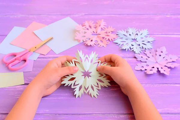 Il bambino ha tagliato un fiocco di neve di carta colorata. Il bambino piccolo tiene un fiocco di neve di carta in mani. Fogli di carta colorati, forbici, fiocco di neve su sfondo di legno — Foto Stock