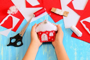 Çocuk keçeli Noel ağacı süslemesi dikmiş. Çocuğun elinde Noel evi süsü var. Mavi ahşap arka planda dikiş aletleri ve malzemeler. Çocuklar için kolay Noel süslemeleri. Çocuk el işi proje fotoğrafı.