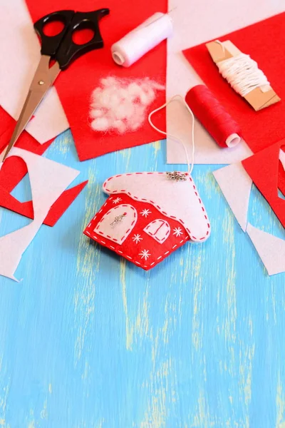 Christmas house ornament sewn from red and white felt, scissors, felt sheets and scraps, needle, thread spool on a blue wooden background with copy space for text. Easy and cheap Christmas DIY — Stock Photo, Image