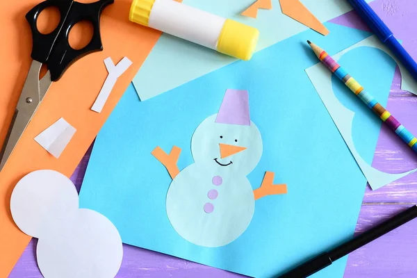 Paper card with snowman applique and text I love winter. Scissors, glue stick, pencil, markers, paper sheets and scraps on wooden background. Simple and fun kids winter crafts. Top view