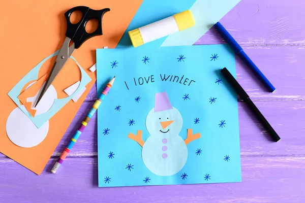 Maken van een kinderen winter papieren kaarten. Stap. Papieren kaart met sneeuwpop collage en tekst die ik hou van winter. Briefpapier en materialen op houten achtergrond. Kinderen winter ambachten concept. Close-up. Bovenaanzicht — Stockfoto