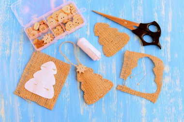 Çuval bezi Noel ağacı süslemesi takılı ahşap yıldız düğmesi. Kağıt ağaç şablonu, iplik, raptiye, makas, mavi arka plandaki tahta düğmeler. Noel dekorasyonu için basit ve ucuz bir el yapımı fikir. Üst görünüm