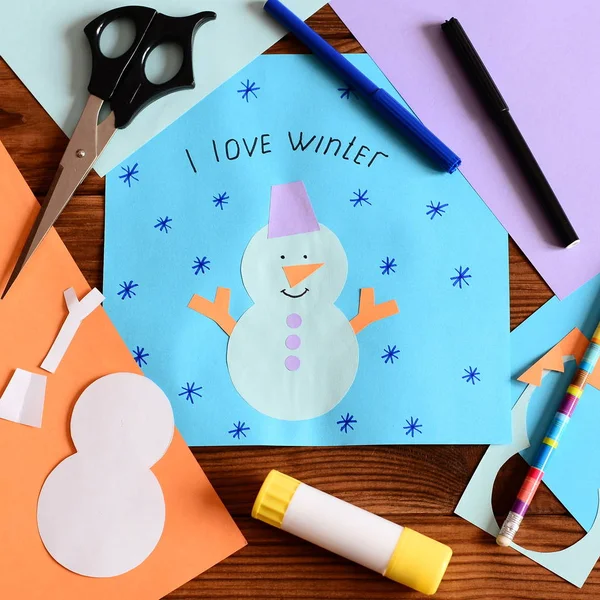 Child made a paper card with a snowman, snowflakes and words I love winter. Craft supplies and paper cutting tools for kids on a wood table. December fine motor activity in kindergarten at home. Children paper crafts diy background. Closeup. Top view — Stock Photo, Image
