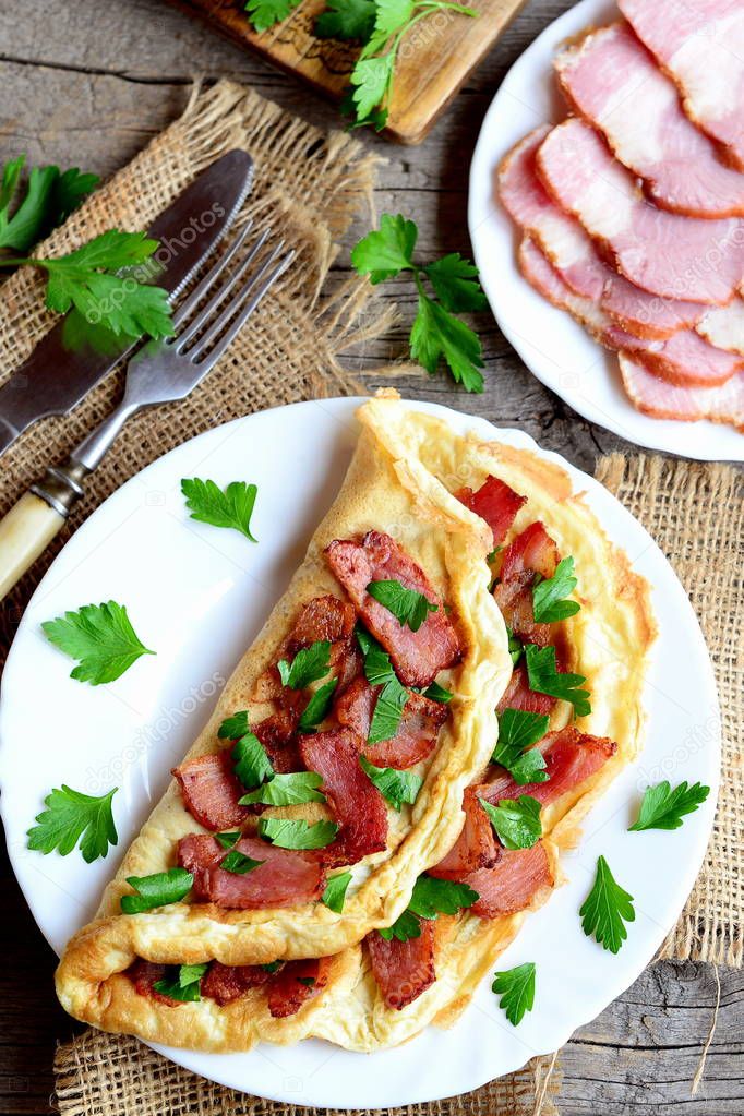 Tasty bacon omelette. Fried omelette stuffed with bacon and parsley for breakfast. Bacon slices on a plate, knife, fork, fresh parsley sprigs on old wooden table. Egg recipe idea. Top view. Closeup