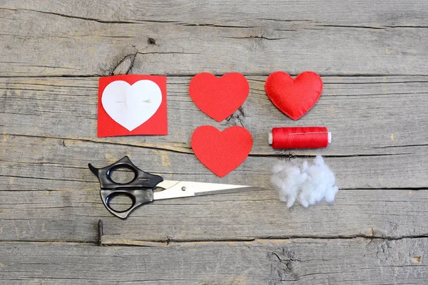 How to hand sew a felt heart for Valentines day. Tutorial. Red felt heart, cut felt pieces in shape of a heart, synthetic filler, paper template, scissors, thread, needle on a wooden table. Top view