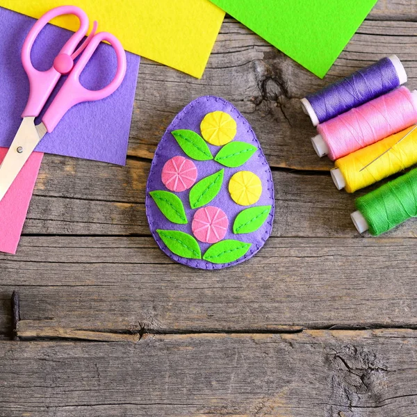 Felt Easter egg decorated with green leaves and colorful flowers. Felt set, threads coils set, scissors on a wooden table. Handmade kids crafts for Easter. Top view — Stock Photo, Image