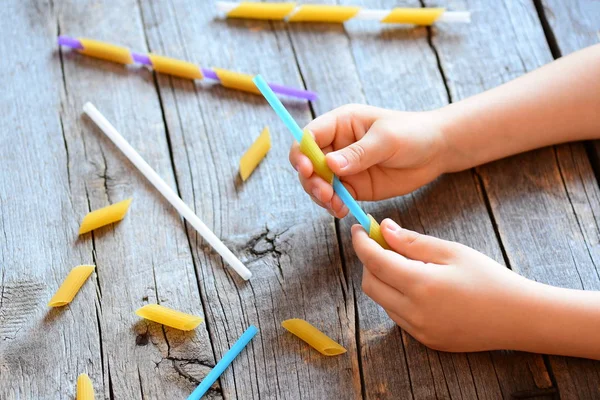 Sviluppo di capacità motorie per bambini. Un bambino teneva in mano una cannuccia e una pasta cruda. Attività creative per bambini. Facile esercizio per migliorare la coordinazione mano-occhio. Sfondo in legno — Foto Stock