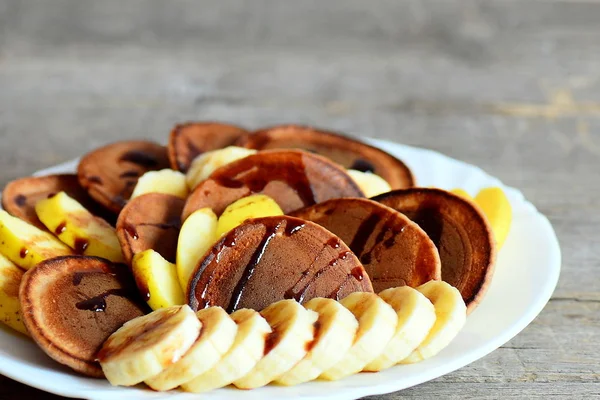 Välsmakande cacao pannkakor. Bakad cacao pannkakor drizzled med sockerkulör, färska skivade bananer och äpplen på en tallrik. Pannkaka dagen frukost och brunch mat idé. Närbild — Stockfoto