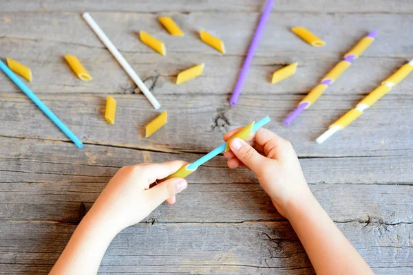Small child holds straw and dried tube pasta in his hands. Child stringing pasta onto straw. Simple task for development of fine motor skills in children