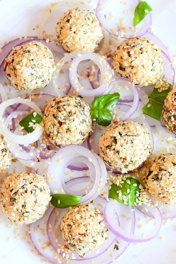 Homemade cream cheese balls on a plate. Cream cheese snacks with dried herbs and toasted sesame seeds served with raw onion rings and fresh basil leaves. Quick cheese snack recipe. Closeup. Top view