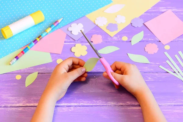 Small child creates a greeting card for mom. Step. Child holds scissors in his hands and cuts a leaf from paper. Materials for paper crafts on a table. Mother's day, birthday, March 8 easy crafts