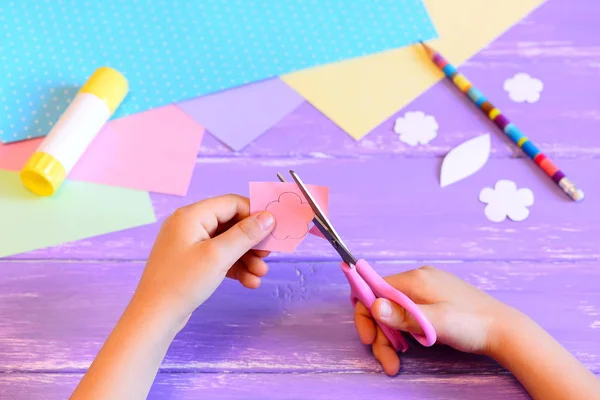 Un niño pequeño crea una tarjeta de felicitación para mamá. ¡Paso! El niño sostiene las tijeras y corta la flor del papel. Materiales para el arte creativo en una mesa de madera. Idea de artesanía para el día de la madre o cumpleaños o el 8 de marzo —  Fotos de Stock