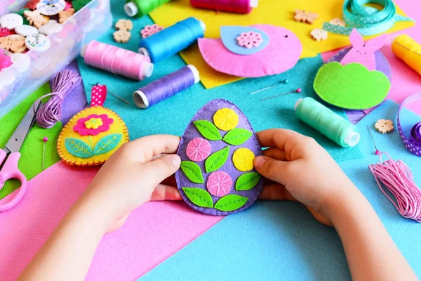 Le gamin tient le décor d'oeuf de Pâques dans ses mains. Petit enfant montre son artisanat de Pâques. Oeufs de Pâques en feutre coloré, décorations de lapins et d'oiseaux, matériaux à coudre et outils sur une table. Bricolage de vacances simple pour les enfants — Photo