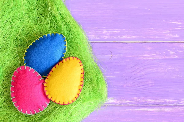 Ροζ, κίτρινο και μπλε τσόχα Πασχαλινά αυγά σε ένα χορτάρι από πράσινη σιζάλ ίνα και σε μωβ ξύλινο φόντο με χώρο αντιγραφής για κείμενο. Ζωντανό πασχαλινό φόντο. Πασχαλινή τέχνη και πανό για παιδιά. Κλείσιμο — Φωτογραφία Αρχείου