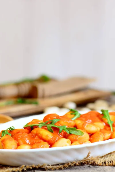 Vegetarian baked beans with carrots, tomatoes and fresh arugula on a plate and on a wooden table. Slow cooked homemade baked beans recipe. Rustic style. Vertical photo — Stock Photo, Image