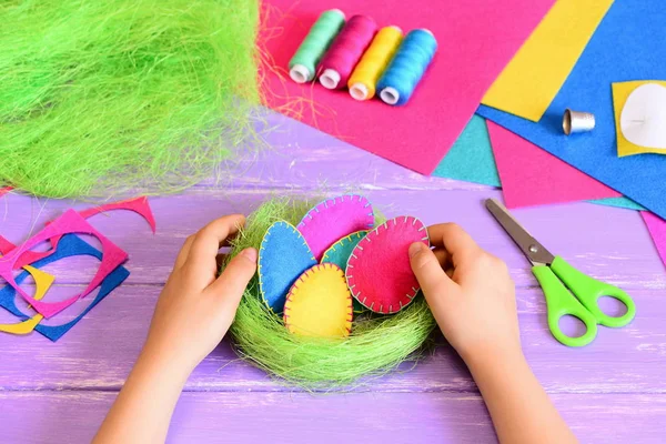 Il bambino piccolo fa la decorazione di Pasqua. Il bambino mette l'uovo di Easter sentito in un cesto. Bellissime uova di Pasqua in feltro e decorazione del nido di sisal, strumenti e materiali artigianali su un tavolo. Regalo di Pasqua facile e bello — Foto Stock