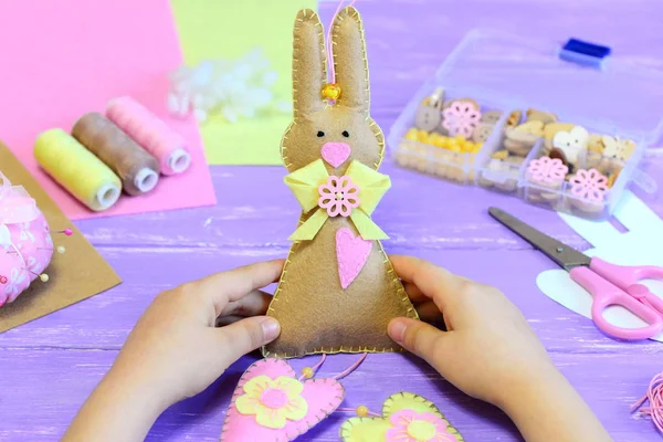 Small child holds a felt rabbit decoration toy in his hands. Child made a felt cute rabbit with hearts for Easter. Tools and materials for children creativity on a wooden table. Simple Easter diy crafts project for kids. Easter holiday concept — Stock Photo, Image