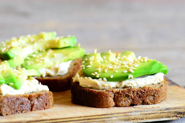 Откройте бутерброды с авокадо и хумусом с кунжутом на деревянной доске. Здоровые веганские сэндвичи. Крупный план — стоковое фото