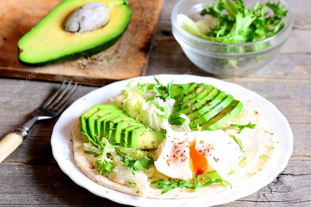 Vegetarian breakfast with a poached egg, avocado slices, cabbage, lettuce mix, tortilla, sauce and spices. Egg and avocado breakfast tortilla recipe. Fork, avocado half on a wooden table