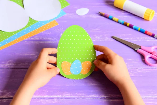 Le petit enfant tient une carte de papier de Pâques entre les mains. Enfant fait carte de vœux créative de Pâques en forme d'oeuf. Matériaux pour l'activité artistique sur une table en bois. Concept artisanal artisanal en papier à la maternelle ou à la maison — Photo