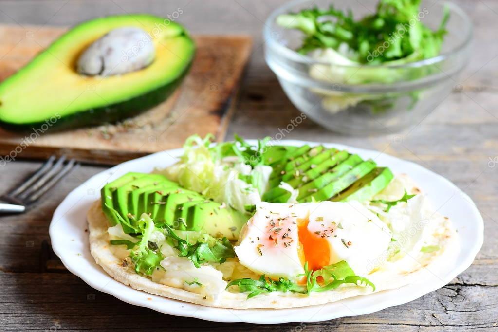 Healthy breakfast concept. Homemade tortilla with a poached egg, avocado slices, napa cabbage, salad mix, sauce and spices on a plate. Avocado half, salad in a glass bowl on a wooden table. Closeup