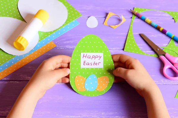 Small child holds a Easter greeting card in hands. Child made a Happy Easter greeting card diy in egg shape. Imagination and creativity for kids development. Easter crafts concept. Stationery on a wooden table — Stock Photo, Image