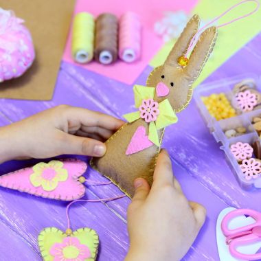 Küçük kız bir keçe Paskalya bunny oyuncak elinde tutar. Kız kalpler keçe bir tavşanın gösterir. El yapımı Paskalya hediyesi. Araç ve Gereçleri çocuklar yaratıcılık ahşap bir masa üzerinde. Çocuklar için eğlenceli el sanatları projesi