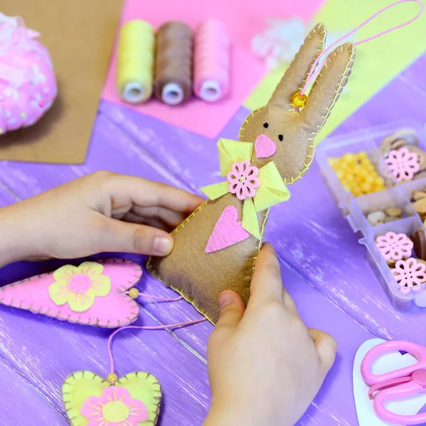 Küçük kız bir keçe Paskalya bunny oyuncak elinde tutar. Kız kalpler keçe bir tavşanın gösterir. El yapımı Paskalya hediyesi. Araç ve Gereçleri çocuklar yaratıcılık ahşap bir masa üzerinde. Çocuklar için eğlenceli el sanatları projesi