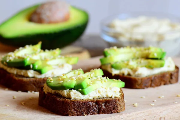 Sandwichs copieux au houmous aux pois chiches et à l'avocat sur une planche de bois, moitié avocat, houmous dans un bol en verre. Sandwichs végétariens cuits avec pain de seigle, tranches d'avocat, houmous de pois chiches et graines de sésame frites — Photo