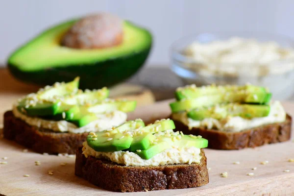 Sandwichs faits maison au houmous et à l'avocat sur une planche en bois, moitié avocat, houmous dans un bol en verre. Sandwichs végétariens ouverts à base de pain de seigle, tranches d'avocat, houmous et graines de sésame frites — Photo