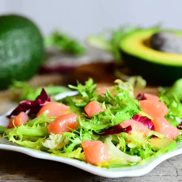 Ensalada de salmón, aguacate y lechuga en un plato. Ensalada dietética con rodajas de salmón ahumado, aguacate fresco y mezcla de hojas de lechuga. La fruta de aguacate y la mitad en una mesa de madera. Primer plano. — Foto de Stock