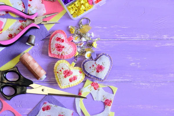 Beautiful hearts and flowers keyring. Homemade felt and fabric keyring or bag charm. Organizer with plastic flowers, scissors, pliers, felt set, fabric, thread. Wooden background with copy space — Stock Photo, Image