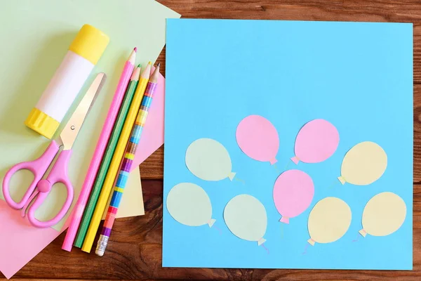 Creating a paper card with balloons. Step. Tutorial for children. Air day or birthday card concept. Card with balloons, scissors, glue stick, colored paper, pencils on a table. Children art activity