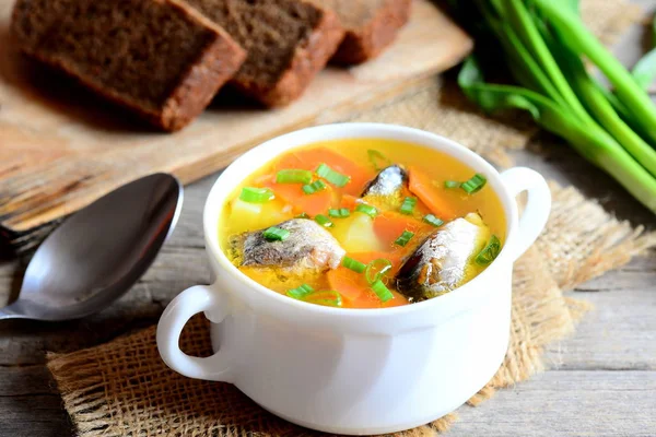 Easy and delicious fish soup recipe. Wholesome fish soup with potatoes, carrots and green onions in a bowl. Rye bread pieces, spoon, fresh green onions on rustic wooden table. Closeup
