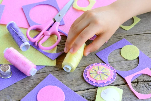 Kindern das Nähen von Hand beibringen. Ein kleines Kind bastelte aus Filzkreisen eine Blume. Kind nimmt Faden in die Hand. Perlenfilzblumen basteln, Bastelmaterial auf einem Holztisch. einfache und schnelle Anwendung — Stockfoto