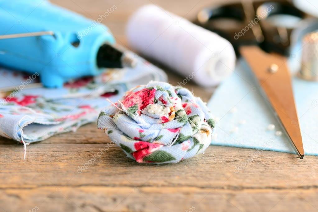 Beautiful flower accessory. Brooch made of fabric with a floral print and embellished with beads, hot glue gun, scissors, thread, fabric, felt on rustic wooden surface. Summer flower accessory crafts