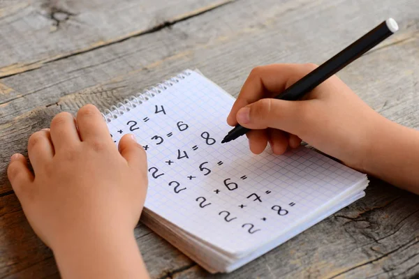 Child resuelve ejemplos matemáticos. Niño sostiene un marcador en su mano y escribe respuestas a ejemplos de multiplicación en un cuaderno. Concepto de tabla de multiplicación de aprendizaje. Educación en el hogar. Niños haciendo deberes. Kid matemáticas estudiar —  Fotos de Stock