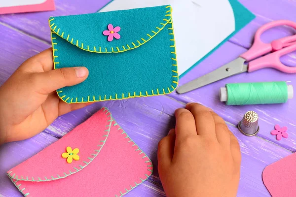 Small child holds a felt purse in his hand. Child shows a felt purse. Handcraft supplies on a wooden table. Funny and easy hand sewing project for kids — Stock Photo, Image