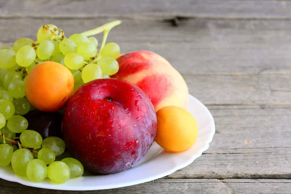 Сырые фрукты на белой тарелке и старинном деревянном фоне. Сладкие персики, сливы, абрикосы, виноградная ветвь. Естественное здоровое питание. Вкусные летние фрукты. Крупный план — стоковое фото