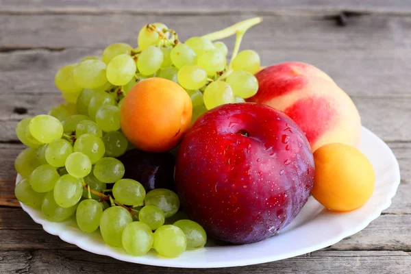 Ассортимент свежих фруктов. Виноград, персик, слива, абрикос на белой тарелке и деревянный фон. Крупный план — стоковое фото