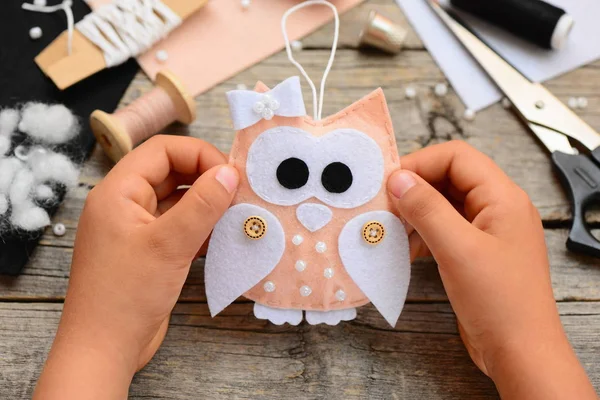 Small child holds a felt Christmas owl ornament in his hands. Sewing supplies and tools on old wooden table. New year decoration idea. Easy kids sewing crafts for Christmas. Closeup — Stock Photo, Image
