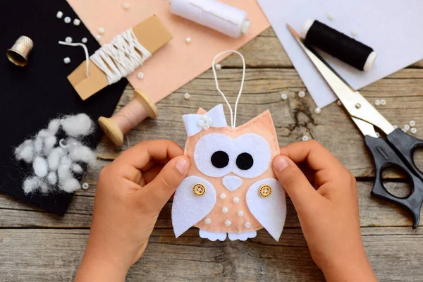 Petite fille montre un décor de sapin de Noël en feutre. Petite fille tient un arbre de Noël hibou décor dans ses mains. Idée de décoration maison pour les enfants. Outils et matériaux artisanaux sur une table en bois. Gros plan — Photo