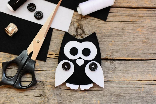 Stuffed felt owl toy, black and white felt sheets, scissors, threads, thimble, black buttons on an wooden background with empty place for text. Beginner sewing projects idea. Top view. Closeup