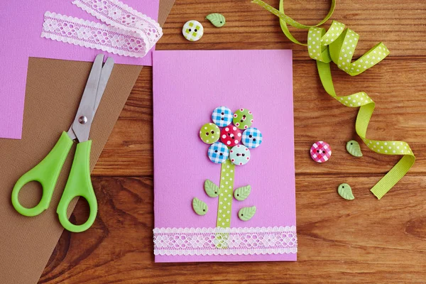 Grußkarte mit einer Blume aus Holzknöpfen, verziert mit Spitze. schöne Papierkartenbasteleien für Mamas Geburtstag oder Muttertag. Werkzeuge und Materialien auf einem Tisch. Kinderarbeitsplätze. Ansicht von oben — Stockfoto