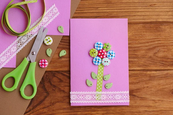 Grußkarte mit einer Blume aus Holzknöpfen, die mit Spitze verziert ist. Geburtstag oder Frauentag 8. März Karte von Kindern für Mutti, Muttertag diy. Werkzeuge und Materialien auf einem Holztisch. Grußkarten können Kinder basteln. Ansicht von oben — Stockfoto