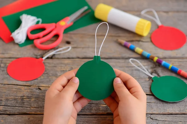 Ребенок сделал красные и зеленые рождественские шарики из картонной бумаги. Ребенок держит рождественский мяч в руках. Шаг. Канцелярские принадлежности на деревянном столе. Легкие елочные игрушки для детей — стоковое фото
